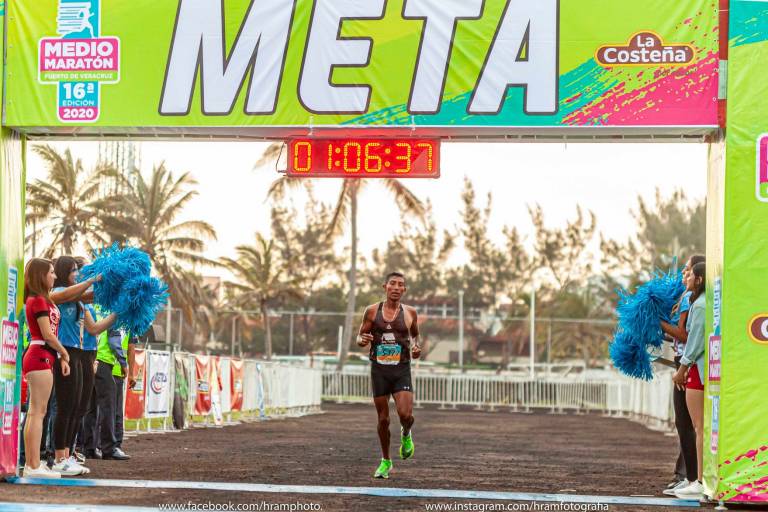 Un corredor cruza la línea de meta en el Medio Maratón de Puerto de Veracruz, con un tiempo de 1:06:37. A ambos lados de la meta, dos animadoras con pompones azules celebran su llegada. En el fondo, se observan palmeras y un cielo despejado, típico de una mañana en Veracruz. El evento, auspiciado por La Costeña, es la 16ª edición del Medio Maratón en 2020.