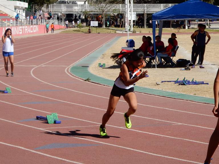 Atleta femenina comenzando una carrera en una pista de atletismo, con espectadores y otros corredores en el fondo.