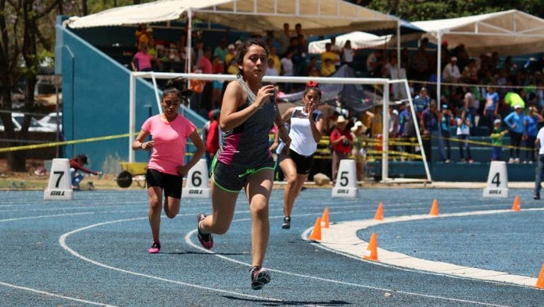 Corredoras compitiendo en una pista de atletismo azul durante una carrera, con espectadores en las gradas al fondo.