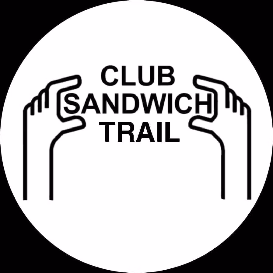 Manos sosteniendo el texto "CLUB SANDWICH TRAIL" en letras negras sobre un fondo blanco circular.